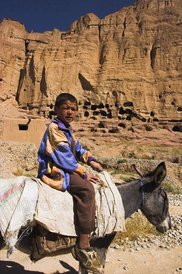 AFGHANISTAN, Bamiyan Province, Bamiyan , Boy on donkey infront of caves in cliffs near empty niche where the famous carved small Budda once stood 180 foot high before being destroyed by the Taliban in 2001