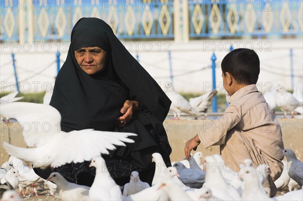 AFGHANISTAN, Mazar-I-Sharif, "Lady wearing burqa and son feeding famous white pigeons at Shrine of Hazrat Ali (who was assissinated in 661) This shrine was built here in 1136 on the orders of Seljuk Sultan Sanjar, destroyed by Genghis Khan and rebuilt by Timurid Sultan Husain Baiqara in 1481, since restored. The shrine is famous for its white pigeons, it is said the site is so holy that if a grey pigeon flies here it will turn white within 40 days"