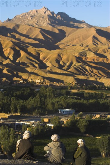 AFGHANISTAN, Bamiyan Province, Bamiyan, Men sit above Bamiyan village as the late afternoon sun glows on mountains near the empty niche where the famous carved Budda once stood (destroyed by the Taliban in 2001)