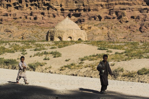 AFGHANISTAN, Bamiyan Province, Bamiyan, Boys walk past tomb near empty niche where the famous carved Budda once stood (destroyed by the Taliban in 2001)