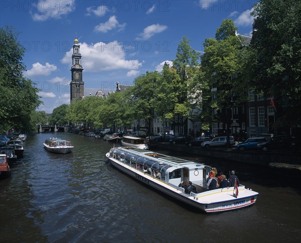 HOLLAND, Noord-Holland, Amsterdam, Pleasure boats on canal lined by trees and parked cars on side streets overlooked by typical architecture partly seen through branches.  Distant bridge and bell tower.