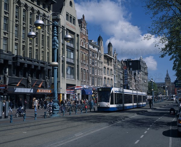 HOLLAND, Noord-Holland, Amsterdam, "Damrak.  Busy street scene, lined with shop, bar and hotel facades.  Crowds of pedestrians, parked bicycles, marked cycle lane and blue and white tram."