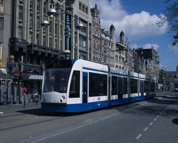 HOLLAND, Noord-Holland, Amsterdam, "Damrak. Blue and white tram on busy street partly obscuring shop, bar and hotel facades. Pedestrians, marked cycle lane and modern street lights."