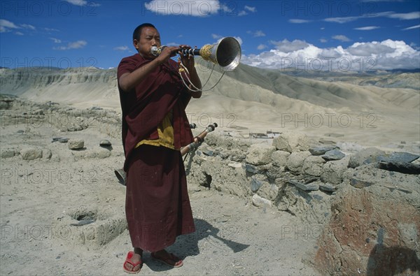 NEPAL, Mustang, Namgyal Gompa, "Tibetan Buddhist monk blowing long, decorated horn on monastery roof to summon the faithful to prayer.  Barren, mountainous landscape behind. Rtnd 2 VKB 15/5/2009"