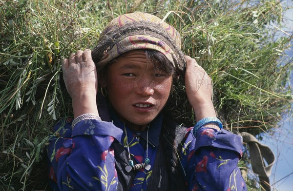 NEPAL, Mustang, Lo Manthang, Portrait of young girl carrying load of harvested grasses supported by strap around her forehead.