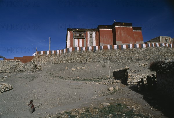 NEPAL, Mustang, Tsarang , "Red, grey and white exterior walls of Tsarang Monastery above flat roof adobe buildings with child on path in foreground."