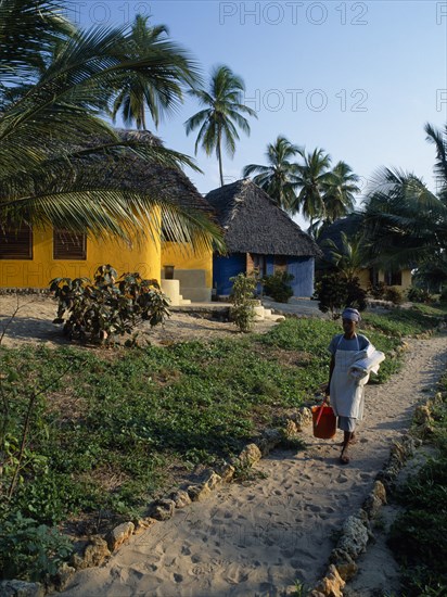 TANZANIA, Pangani, The Tides Hotel. Brightly painted thatched huts amongst palm trees with maid carrying cleaning equipment and fresh towels on path in foreground.