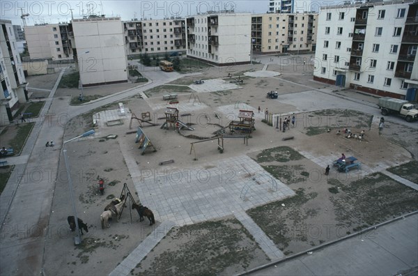 MONGOLIA, Baga Noor, Multi storey housing in coal mining town surrounding playground with children playing and horses tied to street light and swing frame.