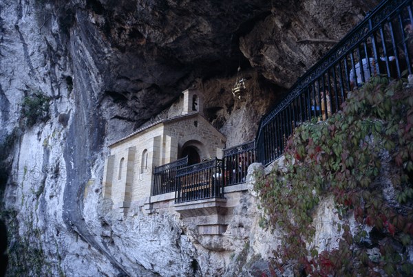 SPAIN, Asturius, Covadonga, Holy Catholic cave shrine in rock face above waterfall and pool.  Contains sarcophagus of the Pelayo who defeated the moors in 718 AD.  People partly seen sitting outside.