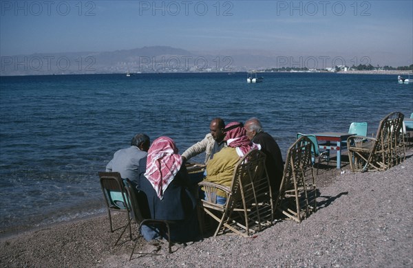 JORDAN, Aqaba, Group of men playing backgammon on shore of beach with Eliat and Israel in distance behind.