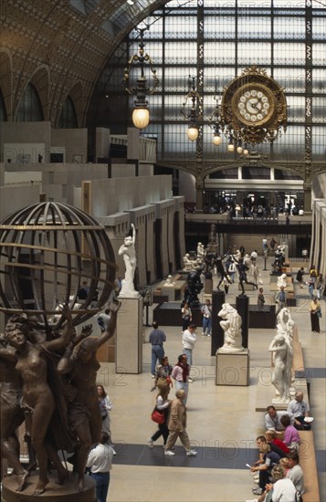 FRANCE, Ile de France, Paris, "Musee d’Orsay interior, visitors walking amongst sculpture inside large gallery space with ornate lighting and clock above."