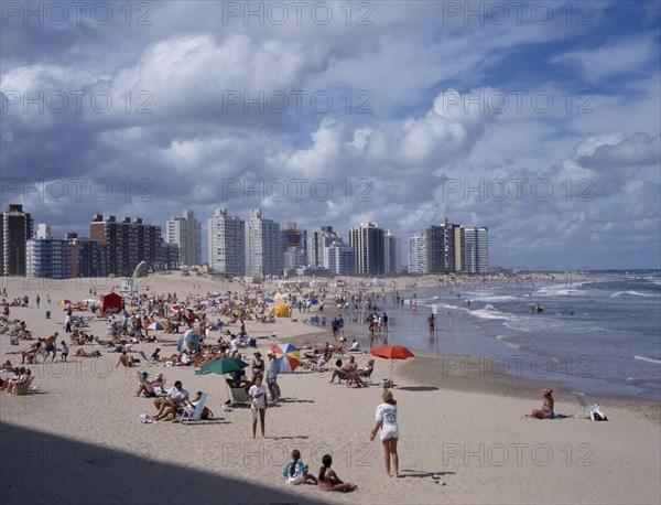 URUGUAY, Punta del Este, "Busy, sandy beach with people sunbathing, playing ball games and in surf.  Overlooked by line of high rise buildings.  "
