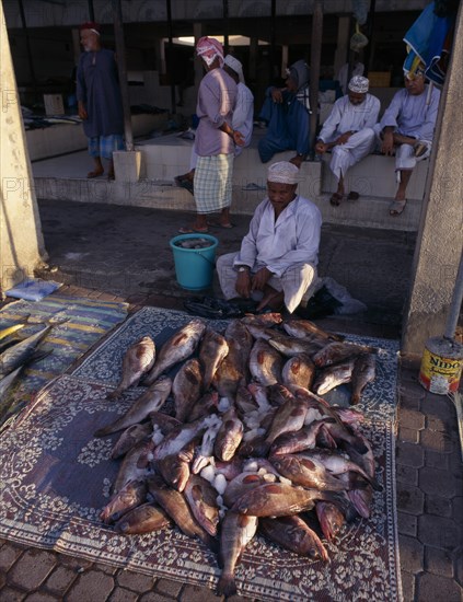 UAE, Oman, Muscat, Mutrah fish market.  Vendor sitting behind display of whole fish scattered with ice laid out on matting on ground in front of him.  Group of men sitting behind.