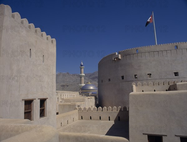 UAE, Oman, Nizwa, Minaret and blue and gold domed roof of mosque seen from fort walls with barren mountainside behind.