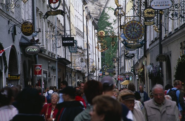 AUSTRIA, Salzburg, Crowds of shoppers on Getreidegasse with cafe and shop signs hanging from decorative metal brackets along length of street above their heads.