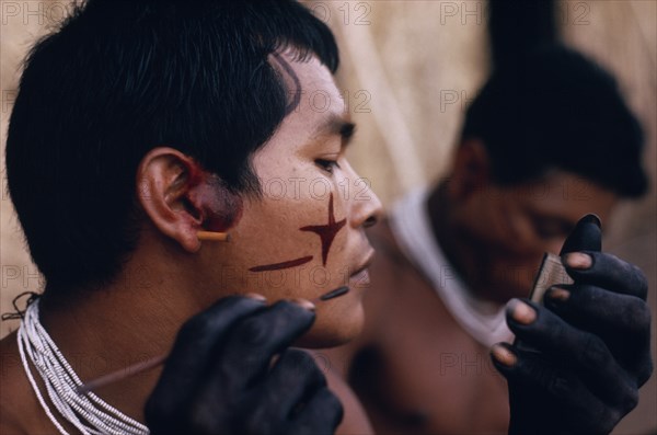 COLOMBIA, North West Amazon, Tukano Indigenous People, Barasana man looking in traded mirror to apply dark red Achiote facial paint with hands and wrists already coloured dark purple from boiled we leaves Tukano sedentary Indian tribe North Western Amazonia body decoration American Colombian Colored Columbia Hispanic Indegent Latin America Latino Male Men Guy South America Tukano