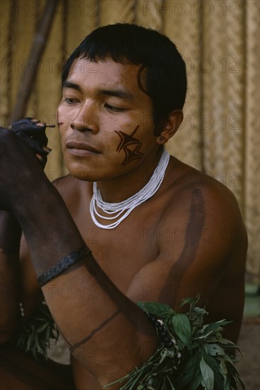 COLOMBIA, North West Amazon, Tukano Indigenous People, Barasana man looking in traded mirror to apply dark red Achiote facial paint with hands already coloured dark purple to wrists from we leaf juice Tukano sedentary Indian tribe North Western Amazonia body decoration American Colombian Colored Columbia Hispanic Indegent Latin America Latino Male Men Guy South America Tukano