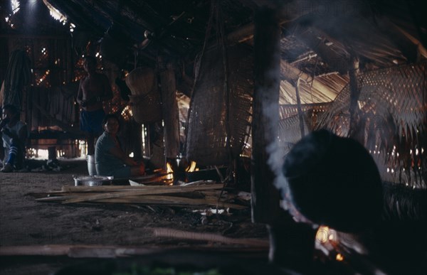COLOMBIA, North West Amazon, Tukano Indigenous People, Makuna maloca or communal tribal home interior with smoke rising from open fire toasting coca leaves to be pounded into fine powder  Woman and two standing men in low light. Tukano  Makuna Indian North Western Amazonia maloca American Colombian Columbia Female Women Girl Lady Hispanic Indegent Latin America Latino Male Man Guy South America Tukano