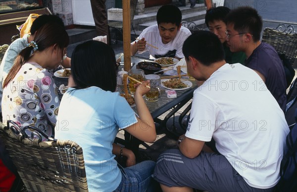 CHINA, Beijing, "Wangfujing shopping street.  Group of young people using chopsticks to eat meal, and drinking at outside table."