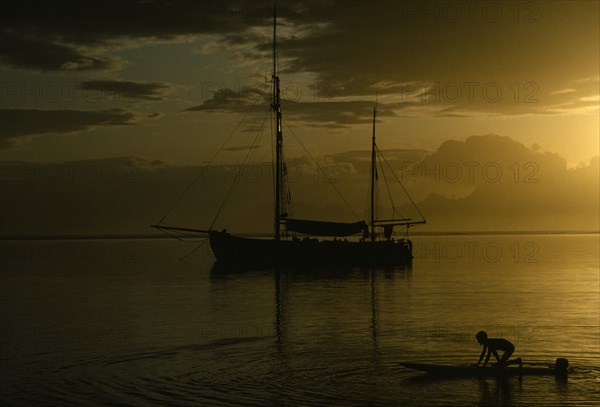 PACIFIC ISLANDS, French Polynesia, Tahiti, Sunset reflected in still expanse of water with silhouetted sail boat at anchor and children playing in the water in foreground.
