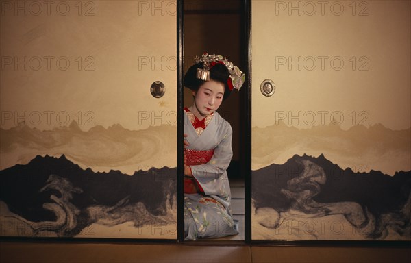JAPAN, Customs, Geisha, "Portrait of Someiyu, a maiko or trainee geisha kneeling in tightly bound kimono between partly open screens with traditional make-up and hair piece in readiness for her evening appointments."