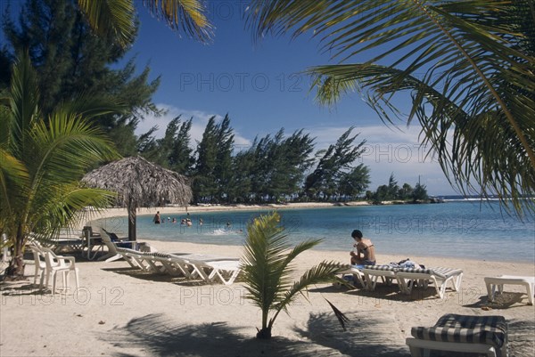 HONDURAS, Roatan Island, Fantasy Island. View through palm tree branches towards  a woman sitting on a sun lounger and people swimming in the bay next to sandy beach