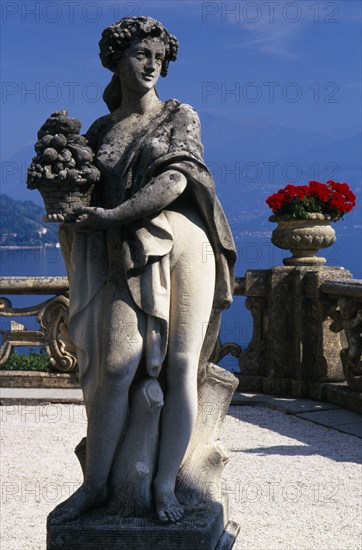 ITALY, Lombardy, Lake Como, Villa Balbianello.  Statue of woman holding basket of fruit with red geraniums in stone vessels on stone balustrade behind and view over lake.