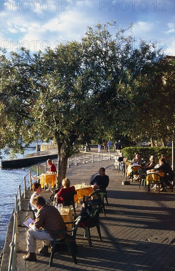 ITALY, Piedmont, Pallanza, Lake Maggiore.  People enjoying lakeside views sitting at cafe tables beneath trees on the esplanade.  Late afternoon sun and long shadows.