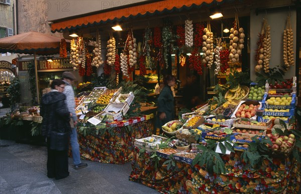 SWITZERLAND, Ticino, Lugano, Member of staff and customers outside fruit and vegetable stall with pavement display and strings of onions and garlic hanging from rack above doorway.