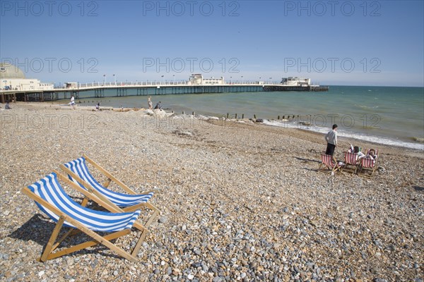 ENGLAND, West Sussex, Worthing, View across beach towards the pier with blue and white deckchairs in the foreground and sunbathers enjoying the sunshine nearby
