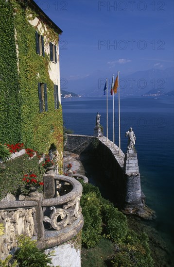 ITALY, Lombardy, Lake Como, "Villa Balbianello.  Part view of ivy and virginia creeper covered facade, stone balcony with pots of red geraniums and wall with statues and flags. View over lake beyond."