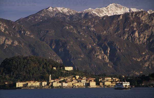 ITALY, Lombardy, Lake Como, Ballagio. View across Lake Como towards distant town of Ballagio situated at foot of tree covered hillside with mountain backdrop. Car ferry crossing lake in foreground.