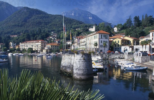 ITALY, Lombardy, Lake Como, Menaggio.  Harbour with moored boats and waterside buildings with tree covered mountain backdrop.