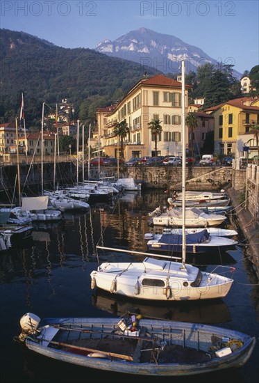 ITALY, Lombardy, Lake Como, Menaggio.  Harbour with moored boats and waterside buildings with tree covered mountain backdrop.