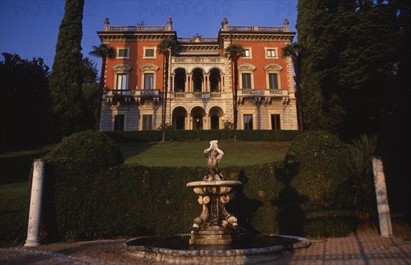 ITALY, Lombardy, Lake Como, "Private villa and formal gardens near Tremezzo, stone fountain in immediate foreground with lawn enclosed by hedge and grand facade of house behind."