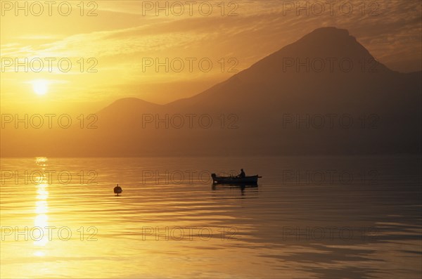 ITALY, Lake Garda, Fishing boat on lake at sunset in golden light with mountain peak in haze of sunshine behind.