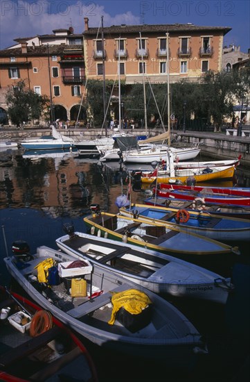 ITALY, Veneto, Lake Garda, Torre di Benaco.  Waterside buildings overlooking harbour with moored boats reflected in water in the foreground.