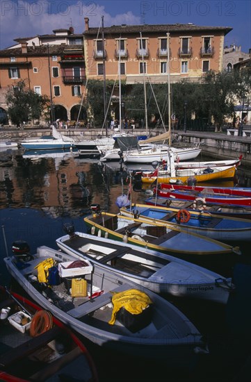 ITALY, Veneto, Lake Garda, Torre di Benaco. Waterside buildings overlooking harbour with moored boats reflected in water in the foreground.