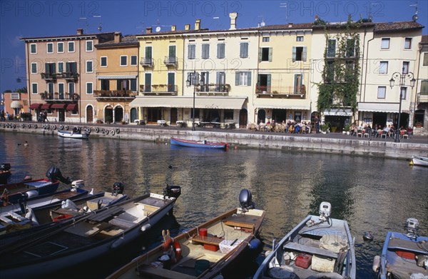 ITALY, Veneto, Lake Garda, Bardolino. Harbour scene with people at outside tables of waterside cafes and line of moored boats in foreground.  Pastel coloured building facades reflected in the water.