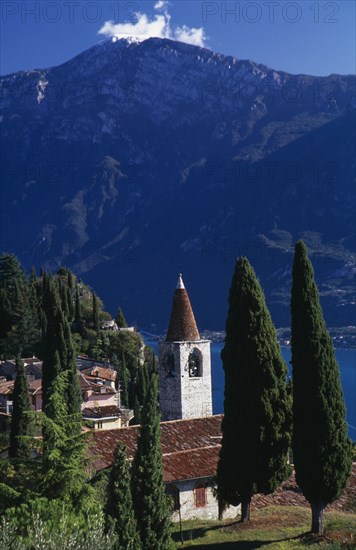 ITALY, Lombardy, Lake Garda, "Pieve.  View of tiled rooftops, church and bell tower of village overlooking Western shore of Lake Garda.  Cypress trees in foreground and mountain backdrop."