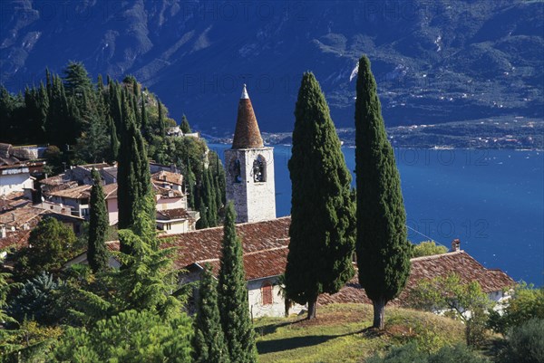 ITALY, Lombardy, Lake Garda, "Pieve.  View of tiled rooftops, church and bell tower of village overlooking Western shore of Lake Garda.  Cypress trees in foreground and mountain backdrop."