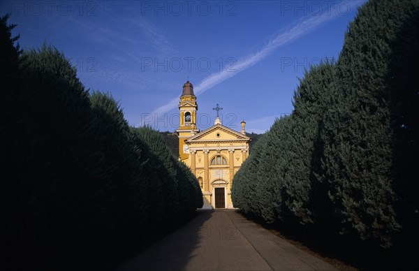 ITALY, Lombardy, Yellow and white exterior facade of small church with bell / clock tower at side with tree lined road leading to entrance.