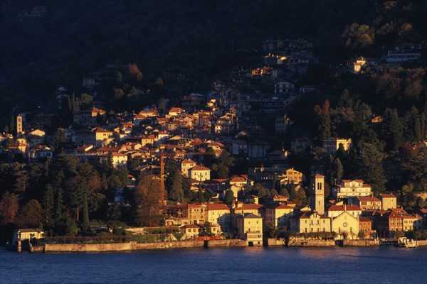 ITALY, Lombardy, Lake Como, Torno.  View across Lake Como towards town spread across hillside in golden sunlight.