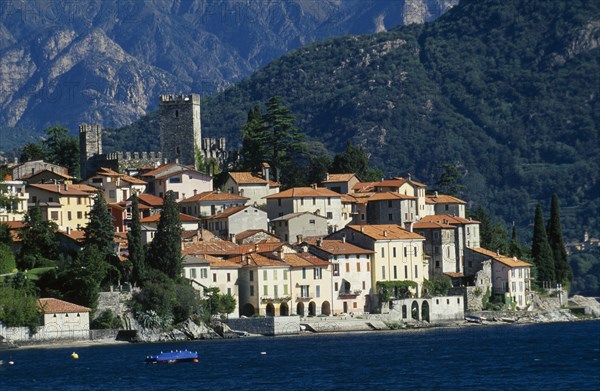 ITALY, Lombardy, Lake Como, Rezzonico. View across lake to red tiled rooftops of village houses with castle tower and walls behind.