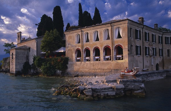 ITALY, Lombardy, Lake Garda , Punta San Vigilio.  Villa on lake shore with couple on sun loungers on raised stone pathway at side.