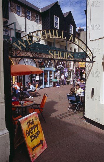 ENGLAND, East Sussex, Hastings, Westhill Arcade. Gate leading to shops with people sitting at cafe tables. A sign advertising The Happy Shop in the foreground.