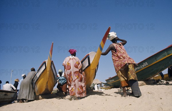 SENEGAL, Soumbedioune, Women wearing brightly coloured traditionally patterned clothing on beach with men and children standing beside painted fishing boats.