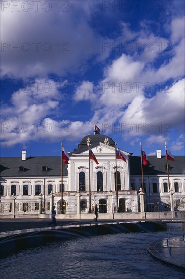 SLOVAKIA, Bratislava, "Presidential Palace, part view of exterior facade with flags.  Circular fountain in the foreground, passing people and Presidential guard either side of entrance."
