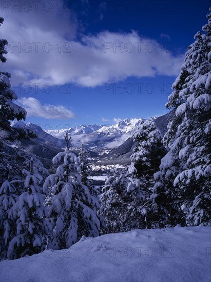 FRANCE, Provence-Cote d’Azur, Hautes-Alpes, View west from Col de Montgenevre over snow covered trees towards Ecrin National Peak and mountain peaks.  View part framed by trees in foreground.