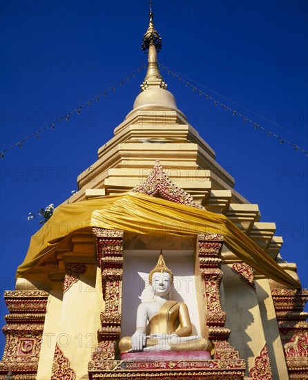 THAILAND, North, Chiang Mai, Seated Buddha statue in decorated recess below gold chedi wrapped in golden orange silk and hung with lights.